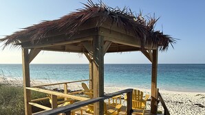 Un kiosque de plage en bois, doté d’un toit de chaume, de chaises Adirondack jaunes et d’une terrasse en bois menant à la plage.
