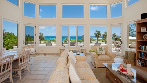 A spacious living room with a large sofa, dining table, and chairs. It features large windows with a view of the ocean and palm trees. - Endless Summer 5 Bedroom Home (Double Bay)