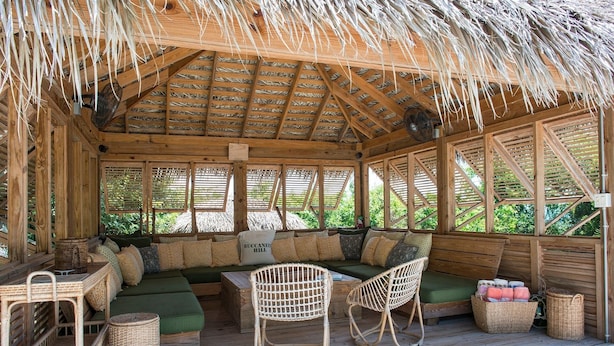A wooden cabin with a thatched roof, a green sofa, wicker chairs, and a wooden table. - Buccaneer Hill Cottage 1 Bedroom Cottage (Governor's Harbour)