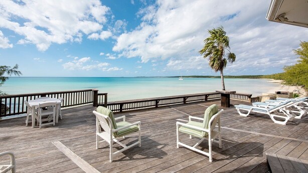 A wooden deck with white chairs and a table, overlooking a beach with clear turquoise water and a sailboat in the distance. - Sand Dollar At Ten Bay Beach 4 Bedroom Home (South Palmetto Point)