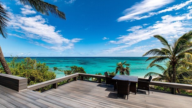Une terrasse en bois avec une table et des chaises, donnant sur une mer turquoise sous un ciel dégagé.