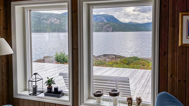 Ein Zimmer mit großen Fenstern, die auf einen See und Berge blicken, Holzboden und Blick auf eine Terrasse mit Gartenmöbeln.