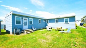 A two-story blue mobile home with a front porch, a small patio with a table and chairs, and a grassy yard. - Bayshore Drive 211 3 Bedroom Home by RedAwning (Ocean City)