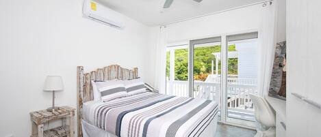 A bedroom with a bed, a ceiling fan, an air conditioning unit, a bedside table with a lamp, and a view of the outdoors through a sliding glass door.