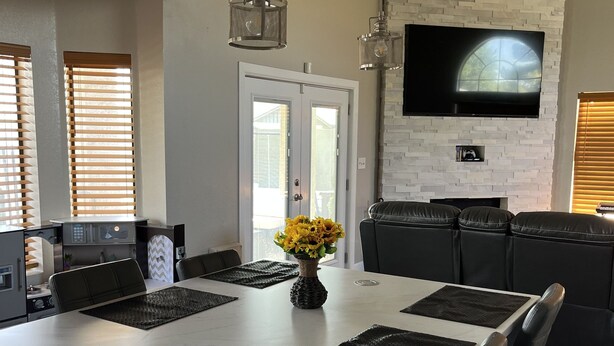 A modern kitchen with a large island, a flat-screen TV mounted on a stone wall, and a dining area with black chairs and a vase of yellow flowers. - Remodeled 2 story home w/ pool 8 minutes from the State Farm Stadium! (Glendale)