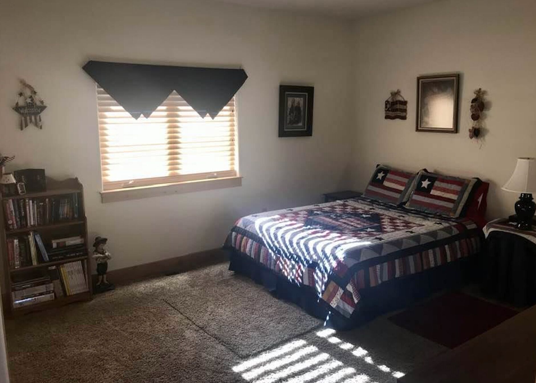Un dormitorio con cama, ventana con persianas, estantería y una mesita de noche con lámpara.