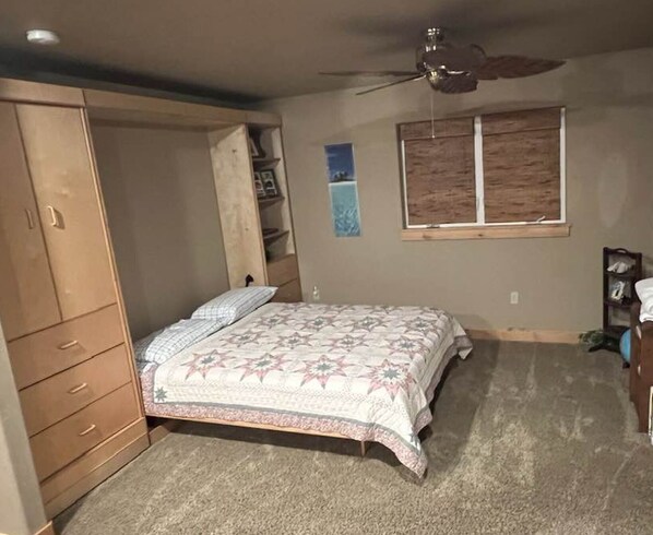 A bedroom with a bed, a wardrobe, a ceiling fan, a window, and a carpeted floor.