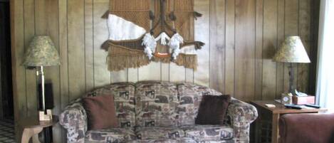 A room with a patterned sofa, a small ottoman, two lamps, and a wall hanging.
