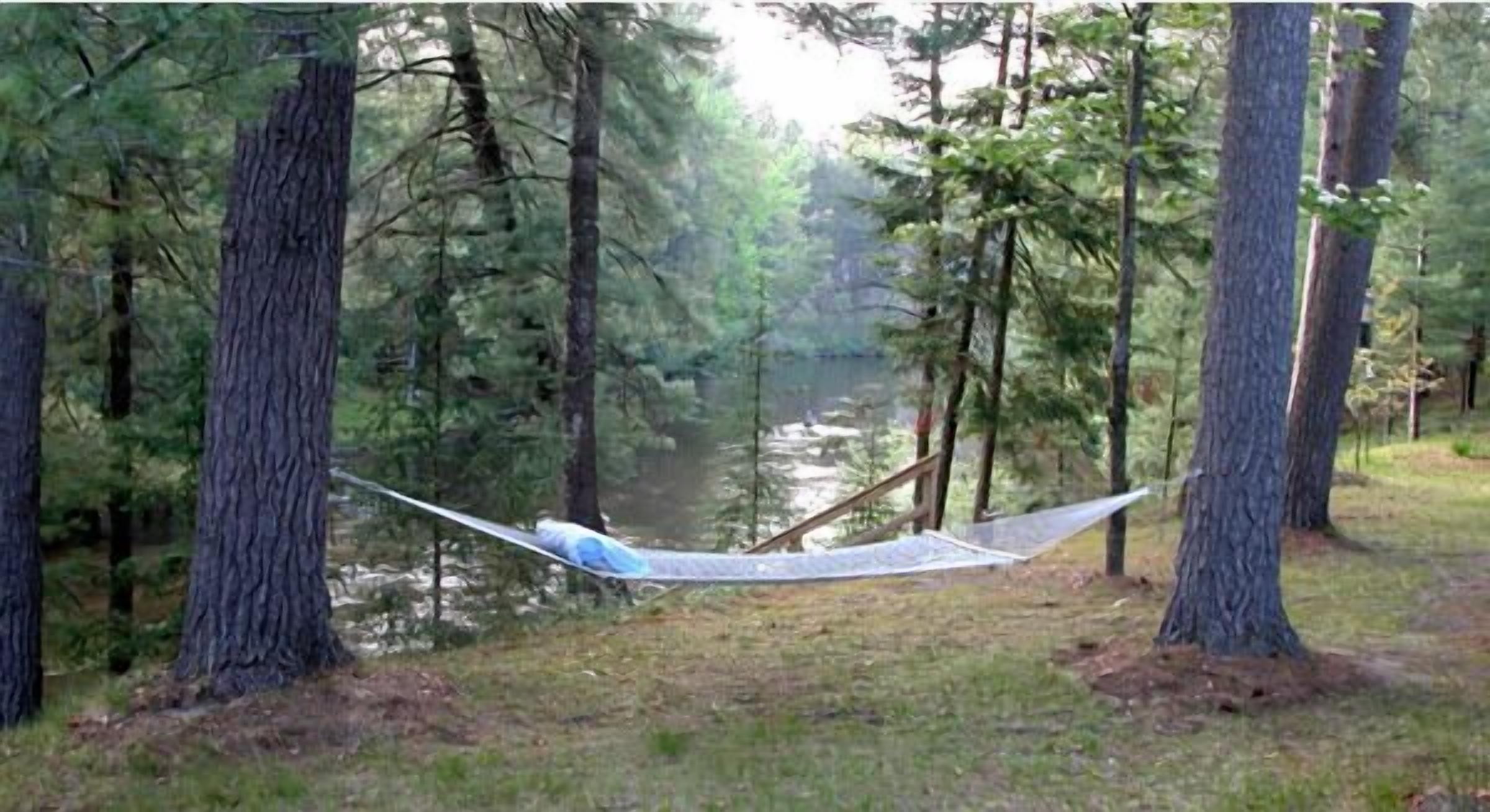 Eine Hängematte zwischen Bäumen in einer bewaldeten Landschaft, im Hintergrund fließt ein Fluss.
