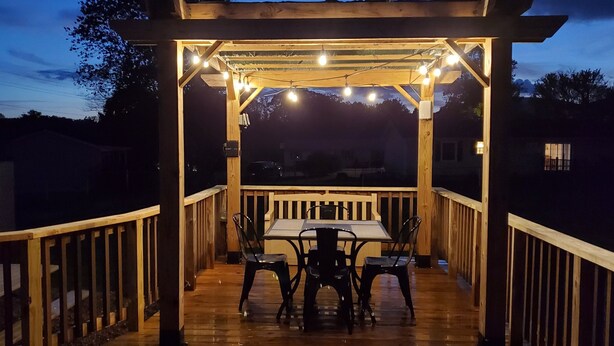 Een houten terras met een tafel, stoelen, lichtjes en een pergola.