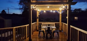 Une terrasse en bois avec une table et des chaises, des guirlandes lumineuses et une structure en forme de pergola.