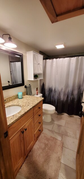 A bathroom with a granite countertop sink, wooden cabinets, a white toilet, and a shower curtain with a forest design. - DuPont Cottage - Bike to DuPont! (Penrose)