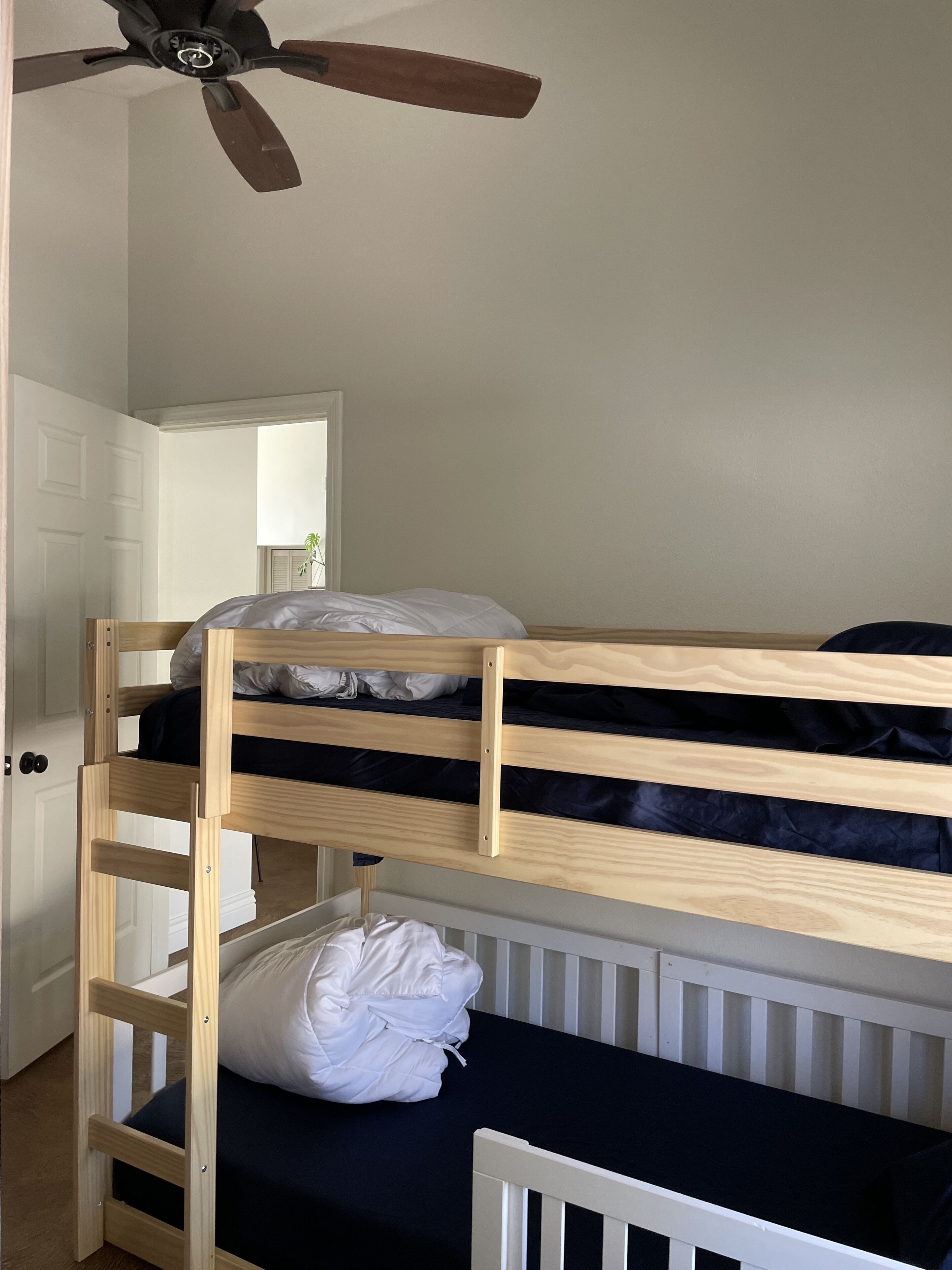 Un lit superposé avec une tête de lit blanche et un berceau blanc en bas. Un ventilateur de plafond est visible au-dessus du lit.