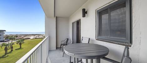 A balcony with a round table and chairs, a view of the beach, and a white wall with a window.
