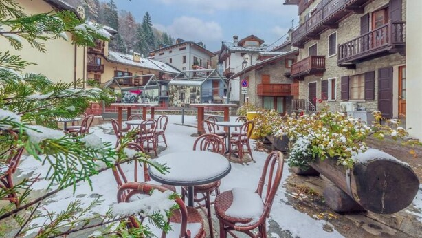 Ein mit Schnee bedeckter Außenbereich mit Sitzgelegenheiten, darunter Korbstühle und -tische, umgeben von Gebäuden mit Balkonen und traditioneller Architektur.