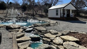 A small white building with a porch, a clear blue sky, and a rocky area with a water feature. - Cozy Hill Cabin 2 (Weatherford)