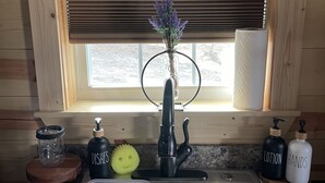 A kitchen with a stainless steel sink, wooden cabinets, a window with blinds, and a vase with flowers on the counter. - Cozy Hill Cabin 2 (Weatherford)