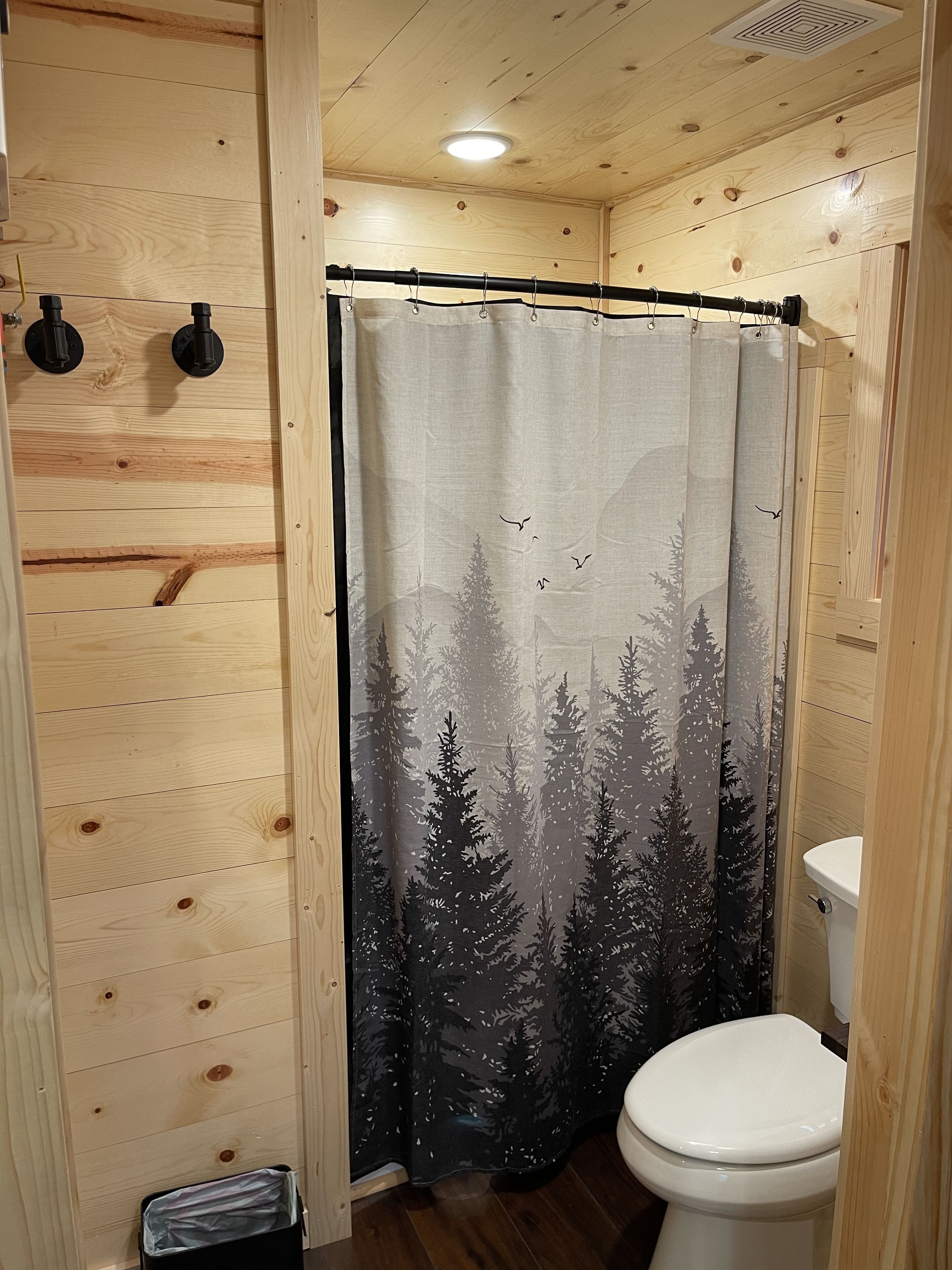 Une salle de bain rustique, avec une cabine en bois, un rideau de douche à motifs de forêt, des toilettes et une petite poubelle.