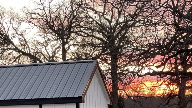 A small cabin with a porch and steps leading up to it, surrounded by trees and a sunset in the background. - Cozy Hill Cabin 2 (Weatherford)