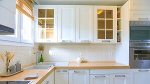 Une cuisine moderne avec des meubles blancs, un plan de travail en bois, un four encastré et une fenêtre avec des stores.