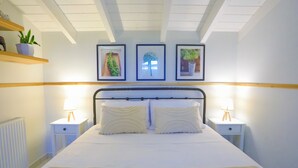 Un lit bien fait, avec du linge de lit blanc et deux oreillers assortis, flanqué de deux tables de chevet avec des lampes, dans une pièce dont le plafond est blanc et qui présente des poutres en bois.
