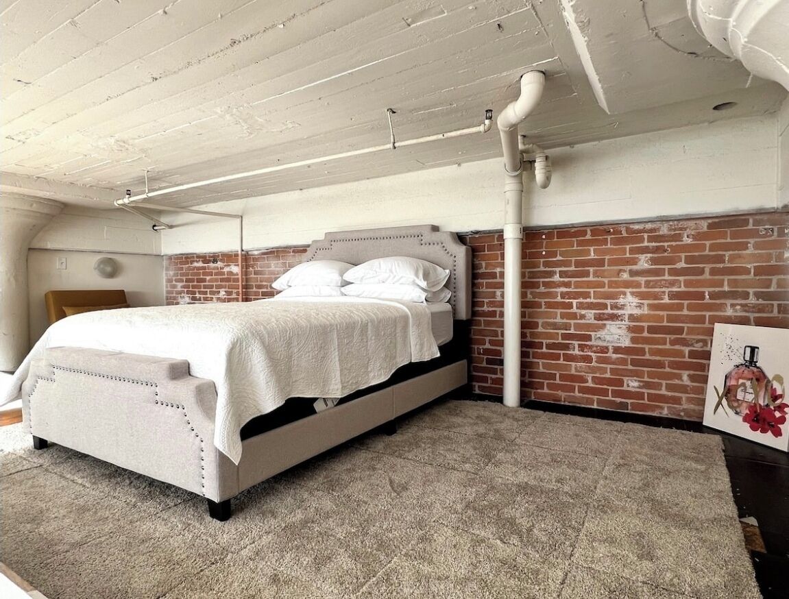 A bedroom with a bed, a brick wall, and exposed ceiling pipes.