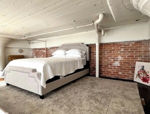 A bedroom with a bed, a brick wall, and exposed ceiling pipes.