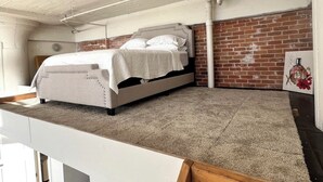 A bedroom with a bed, a white headboard, a brick wall, and a white ceiling with exposed pipes. - Open Loft w/Water Bridge View 2mins to Dwntn (Detroit)