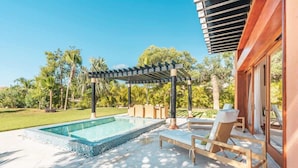 Ein Poolbereich mit einer Pergola, Liegestühlen und freiem Blick auf die umliegende Grünfläche.