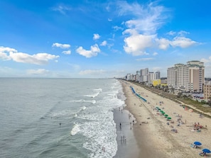 A beach with a long stretch of sand, a pier extending into the water, and several high-rise buildings in the background.