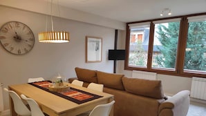 Ein modernes Wohnzimmer mit einem Holzesstisch, weißen Stühlen, einem braunen Sofa, einer großen Wanduhr und einem Fenster mit Blick auf Bäume.