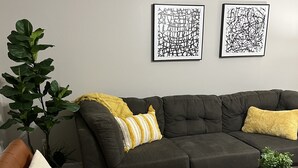 Una sala de estar con un sofá seccional gris oscuro, un sillón de cuero marrón, un tapete estampado y dos obras de arte abstracto en la pared.