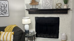 Una sala de estar con chimenea, un sofá negro con una manta amarilla, una mesita de noche con lámpara y una alfombra estampada.