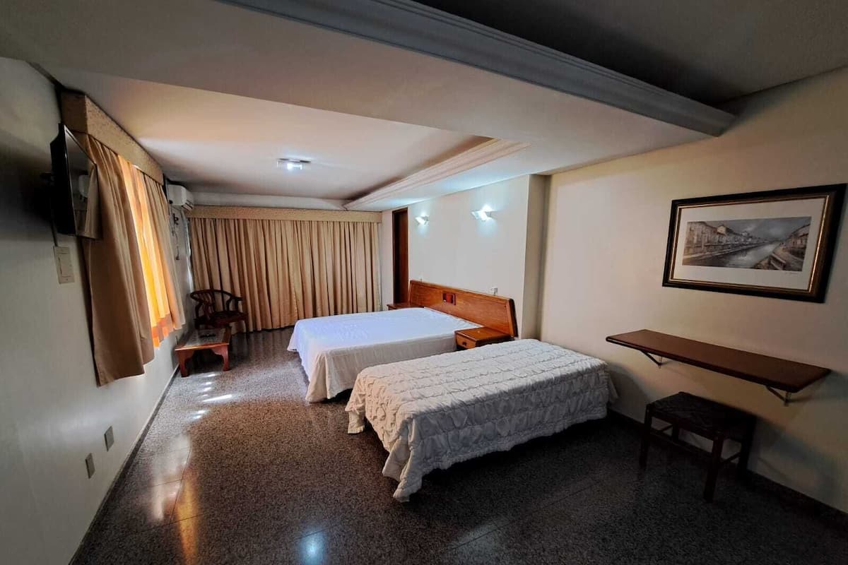 A hotel room with two beds, a desk, a chair, a TV, and a painting on the wall.