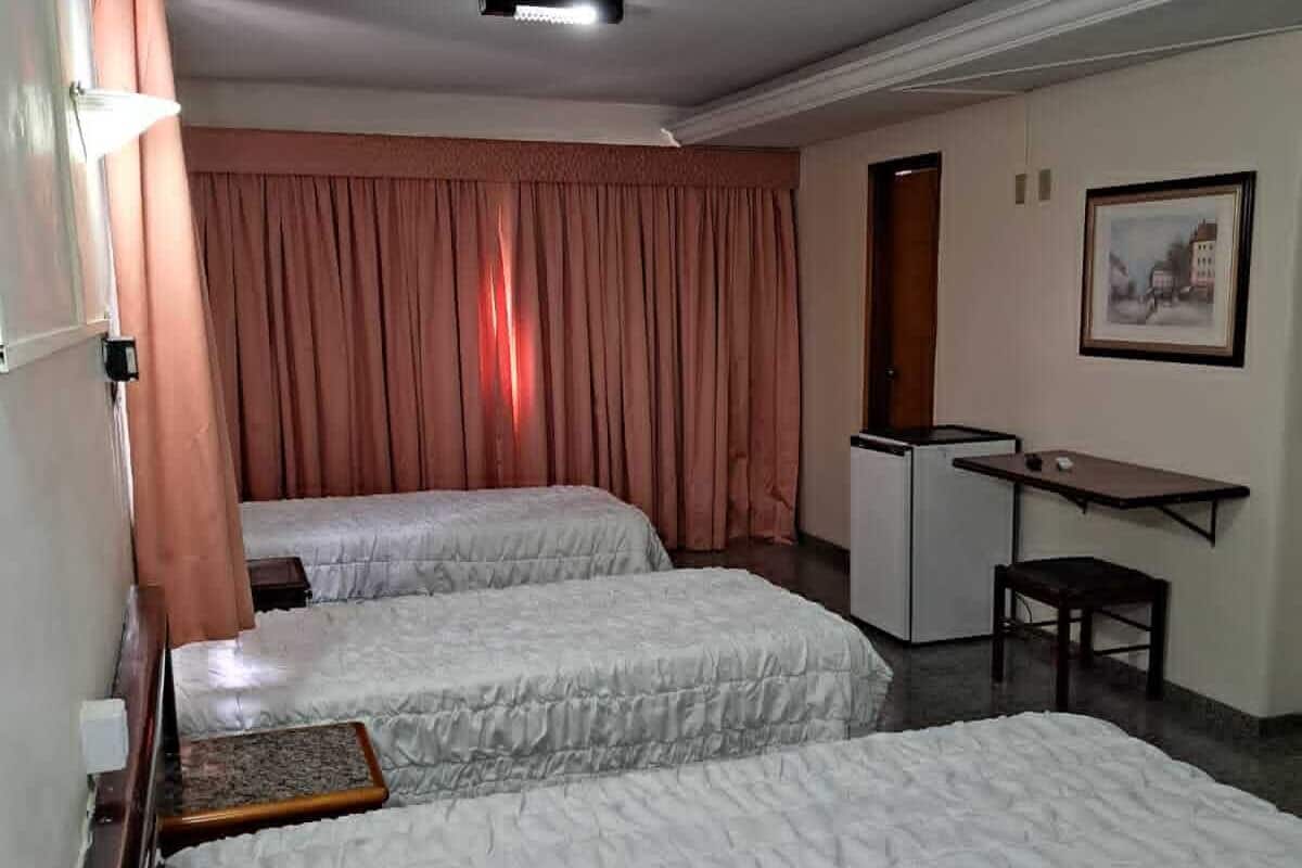 A hotel room with two beds, a desk, a chair, a small table, a painting on the wall, and a window with curtains.