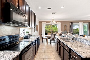 A modern kitchen with dark cabinetry, a large island with a sink, and a dining area with a view. - Ospreys Landing At Solterra Resort 6 Bedroom Home (Davenport)