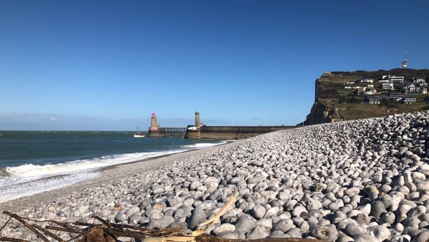 A pebble beach with a lighthouse in the distance and a cliff with buildings on top.