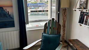 A room with a rocking chair, a view of a harbor, a lamp, and a bookshelf.