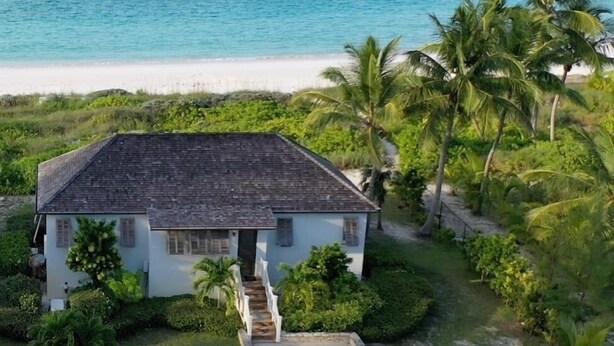 A white house with a dark roof, surrounded by lush greenery and palm trees, located near a beach with clear blue waters. - French Leave South Beach Dogtrot Villa 2 Bedroom Villa (Governor's Harbour)