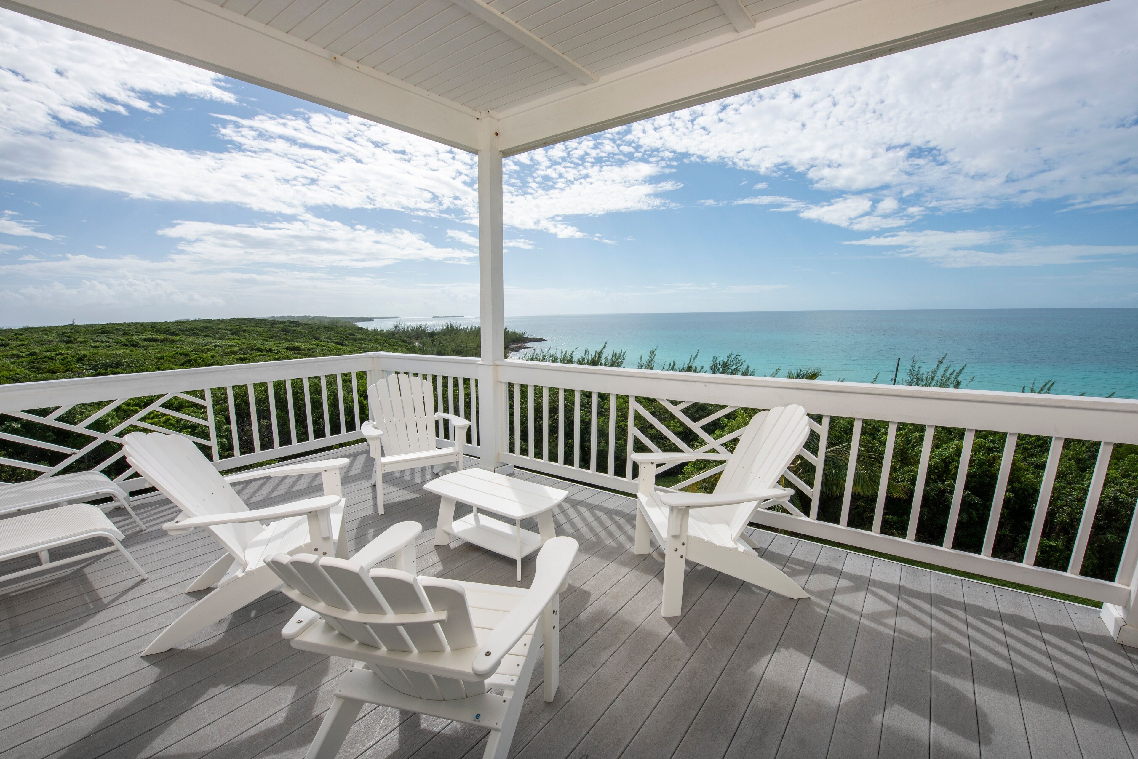 A covered outdoor deck with white Adirondack chairs and a table, overlooking a turquoise ocean and green landscape.