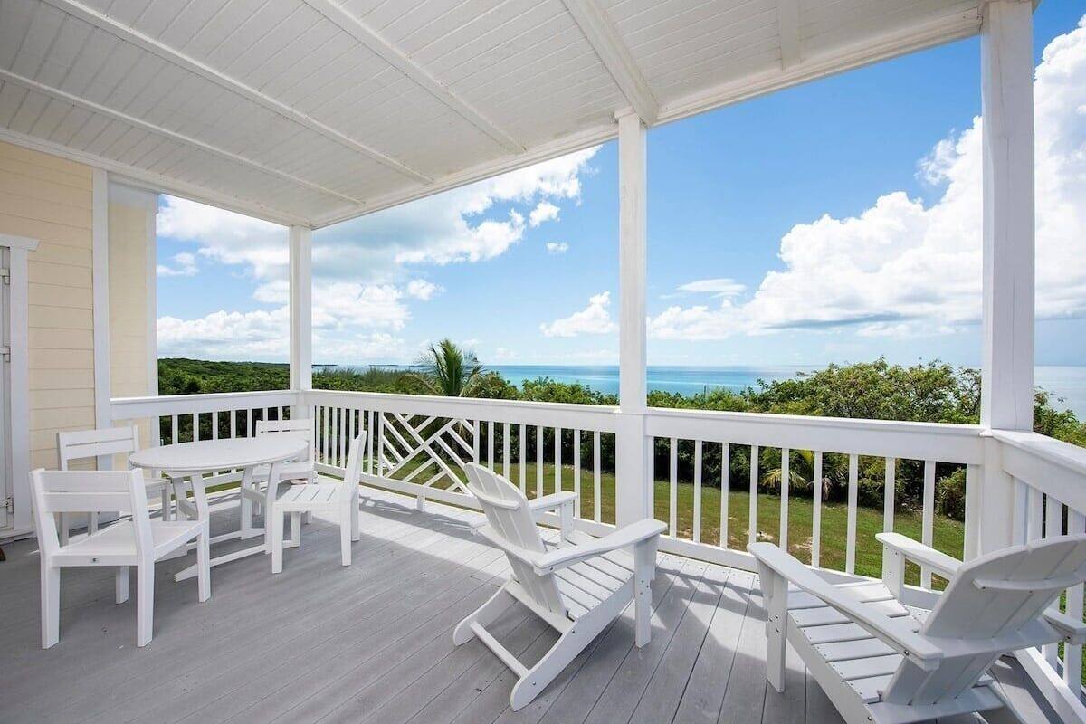 A covered outdoor area with white wooden furniture, including chairs and a table, overlooking a scenic view of the ocean and greenery.
