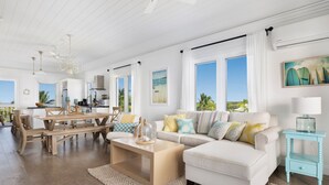 A spacious living room with a white sofa, wooden dining table, and large windows offering a view of the ocean. - Buttonwood Reserve by Eleuthera Vacation Rentals (James Cistern)