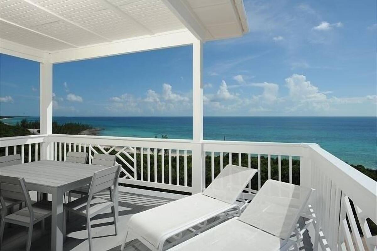 A covered outdoor area with a table and chairs, a white railing, and a view of the ocean.