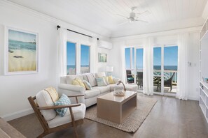 A spacious living room with a white sofa, a wooden chair, a coffee table, and a view of the ocean through large windows. - Buttonwood Reserve by Eleuthera Vacation Rentals (James Cistern)