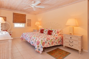 Une chambre à coucher comprenant un lit, deux tables de chevet, un ventilateur de plafond et une fenêtre avec des stores.