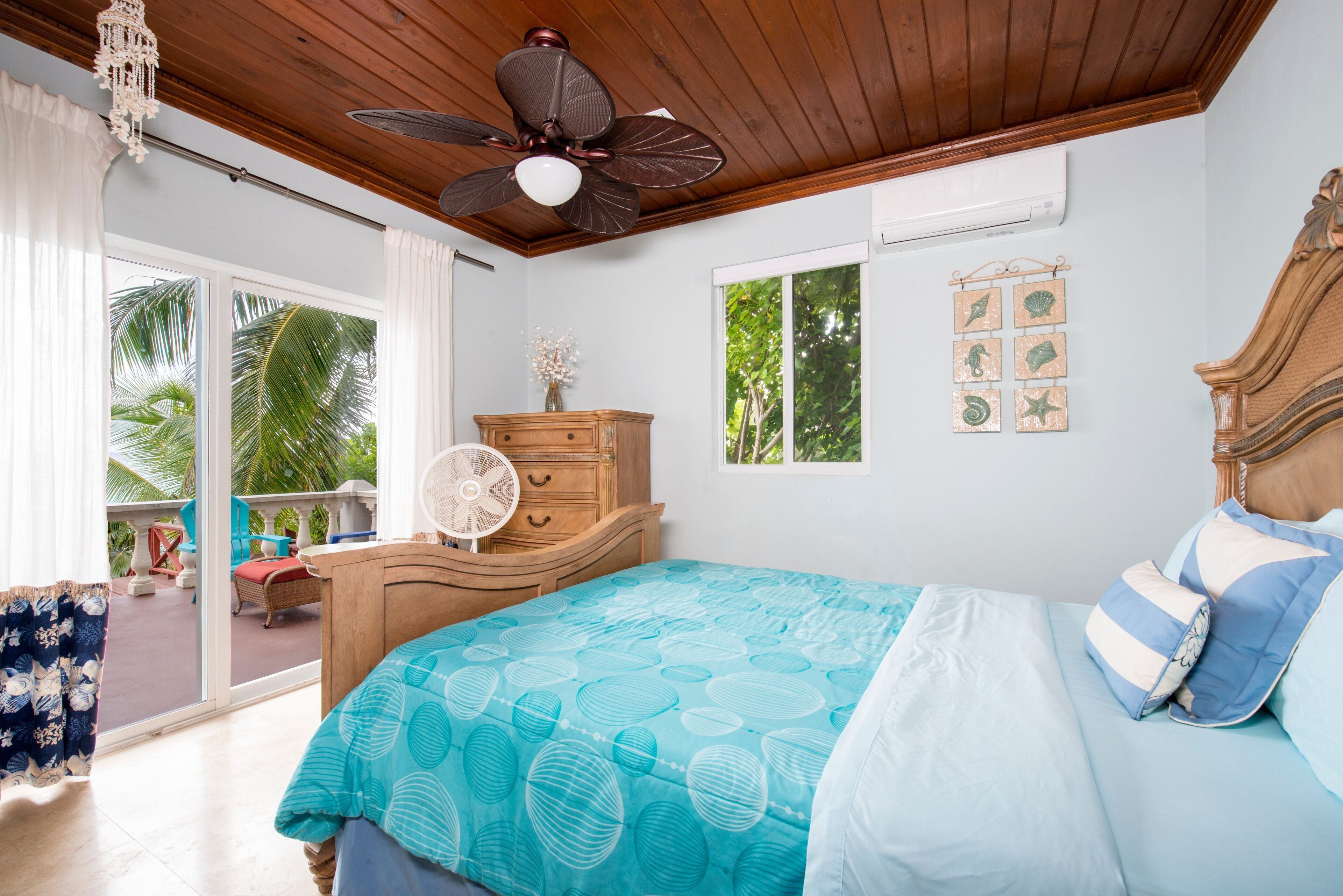 Ein Schlafzimmer mit einem Bett, einer Holzkommode, einem Deckenventilator und Blick auf Palmen durch eine Glastür.
