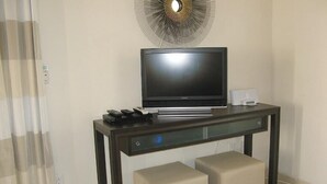 Een moderne hotelkamer met een flatscreen televisie op een donker houten consoles, twee beige poefs en een decoratief kunstwerk boven de televisie.