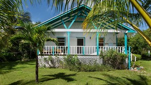 A house with a white exterior and teal accents, surrounded by lush greenery and a clear blue sky. - Orchid Cottage 2 Bedroom Cottage (Governor's Harbour)