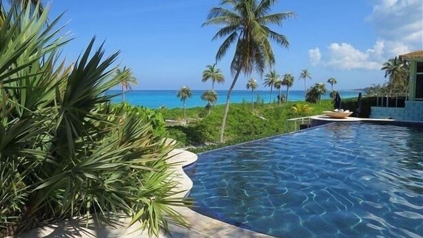 A swimming pool with clear blue water, surrounded by lush greenery and palm trees, overlooking a beach with the ocean in the background. - Orchid Cottage 2 Bedroom Cottage (Governor's Harbour)