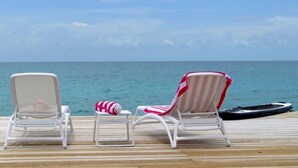 Eine Holzterrasse mit zwei weißen Liegestühlen und einem gestreiften Strandstuhl mit Blick aufs Meer.